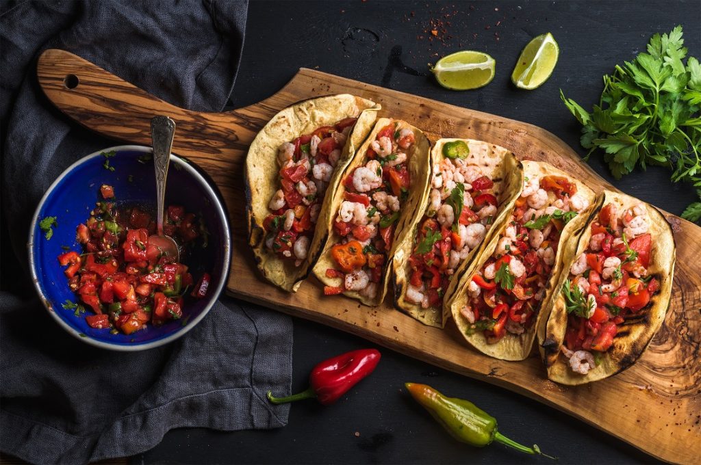 Mexican food- tacos on a wooden slab next to a dish of salsa surrounded by garnishes