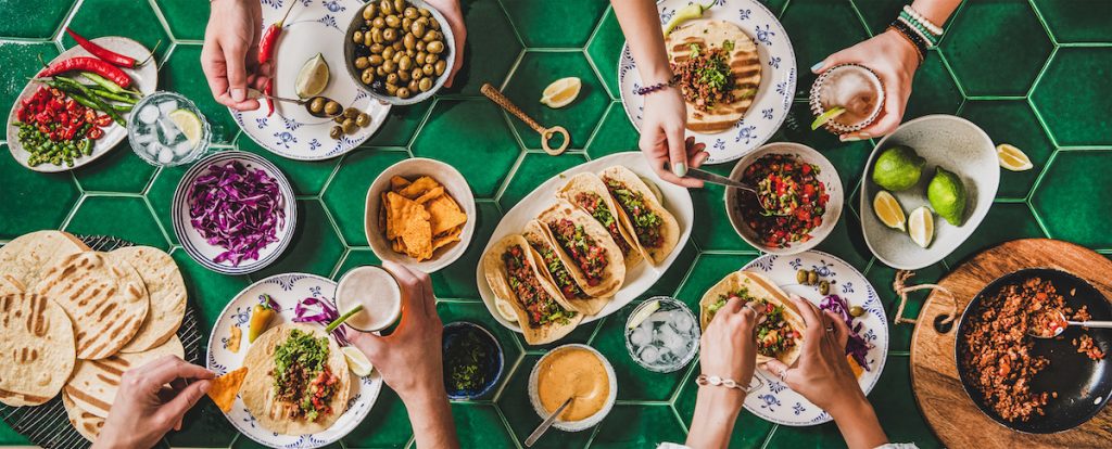 Family or friends home taco party. Flat-lay of Mexican traditional dishes Tacos with beef meat, corn tortillas , tomato salsa and peoples hands over green background, top view. Mexican cuisine concept